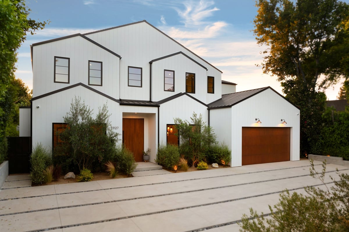 Modern white house with wooden accents and a landscaped driveway under a clear sky, surrounded by greenery.