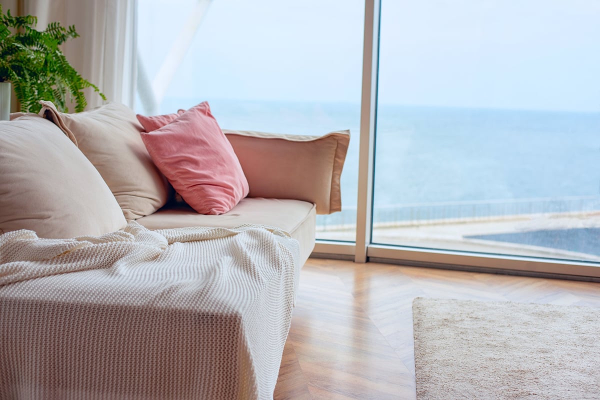 A comfortable beige sofa with pink and cream cushions and a textured throw blanket is placed near a large window with a serene ocean backdrop