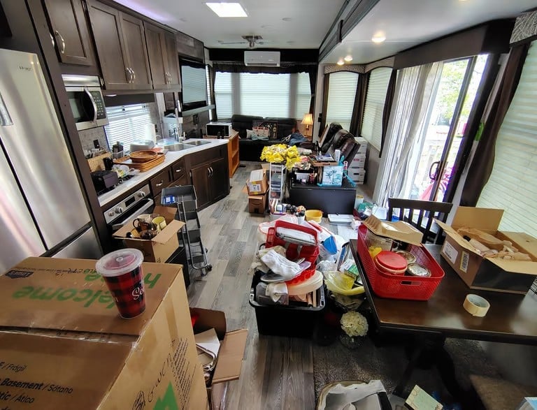 Interior of a cluttered RV with kitchen on left, dining area, and living space with windows, boxes and belongings scattered throughout.