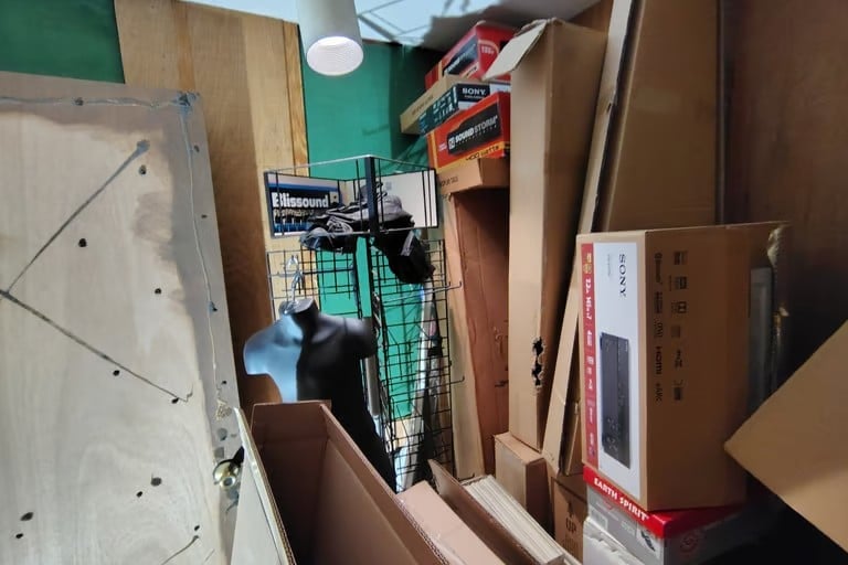 Black cat stands inside a wire storage cage surrounded by cardboard boxes and storage items in a cluttered room