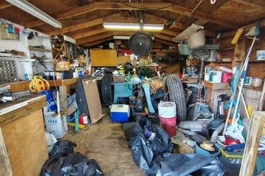 Cluttered garage interior with wooden workbench, scattered tools, storage boxes, and miscellaneous items on shelves and floor