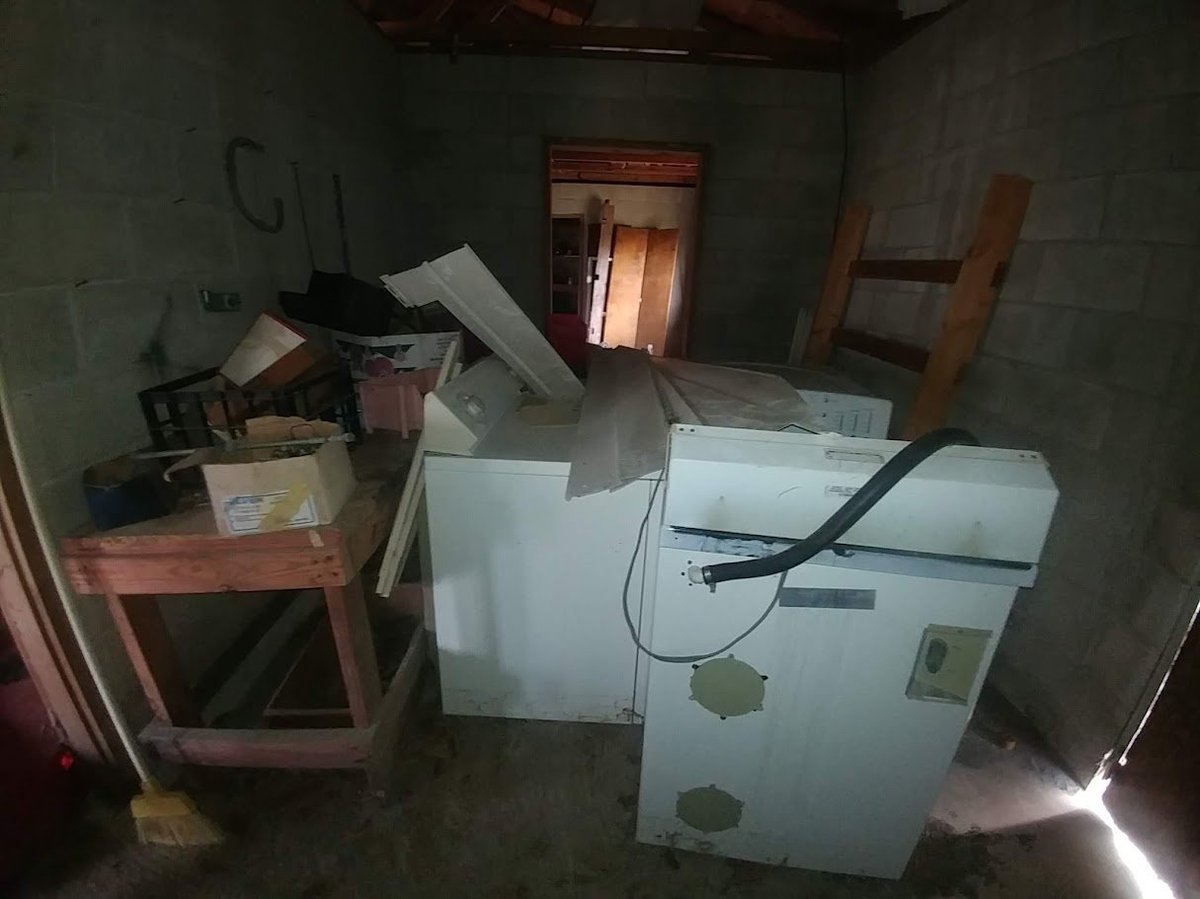 Abandoned room with white appliances, scattered boxes and debris on floor, wooden furniture against walls.