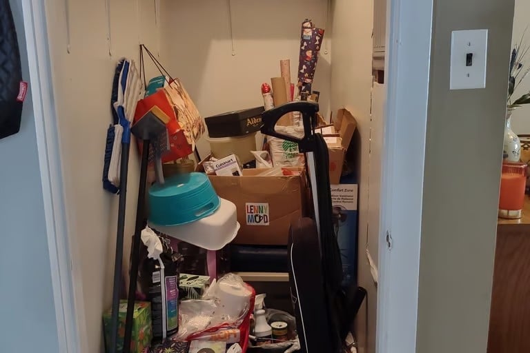 Cluttered closet filled with various items including cleaning supplies, storage boxes, a blue hat, and household goods stacked on shelves