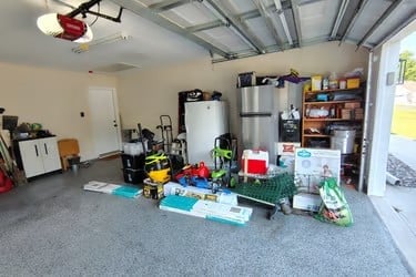 Cluttered residential garage with storage shelves, refrigerator, tools, and various items organized on the concrete floor