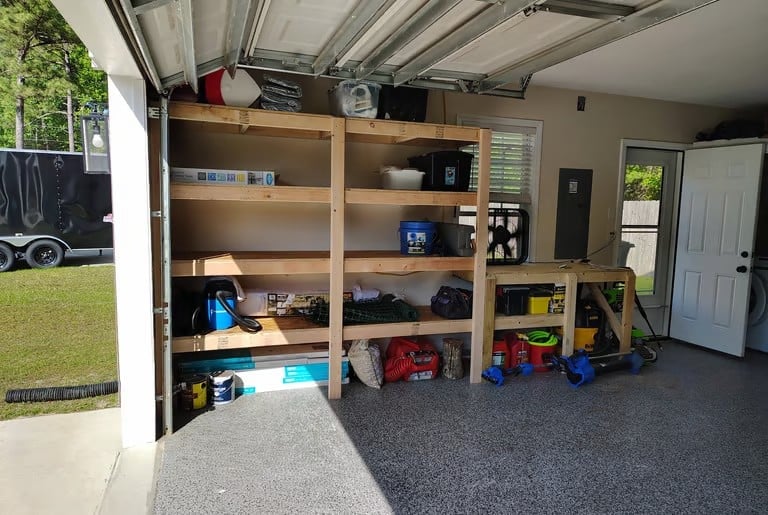Organized garage storage with wooden shelving unit filled with tools, equipment, and household items. Concrete flooring visible with open garage door leading to yard.