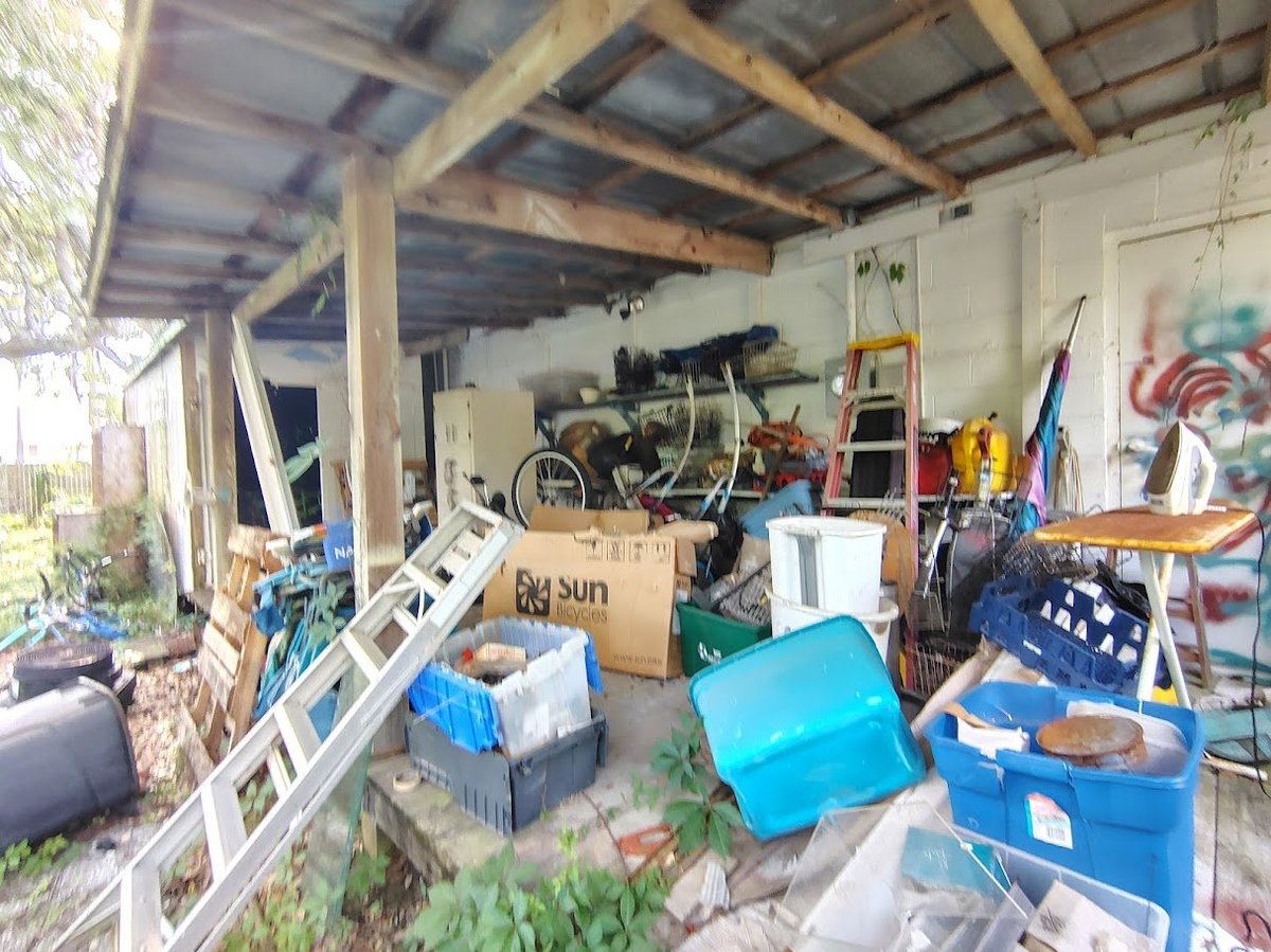 Cluttered garage workshop with wooden beam ceiling, blue storage bins, tools, bicycle, and various equipment scattered throughout.