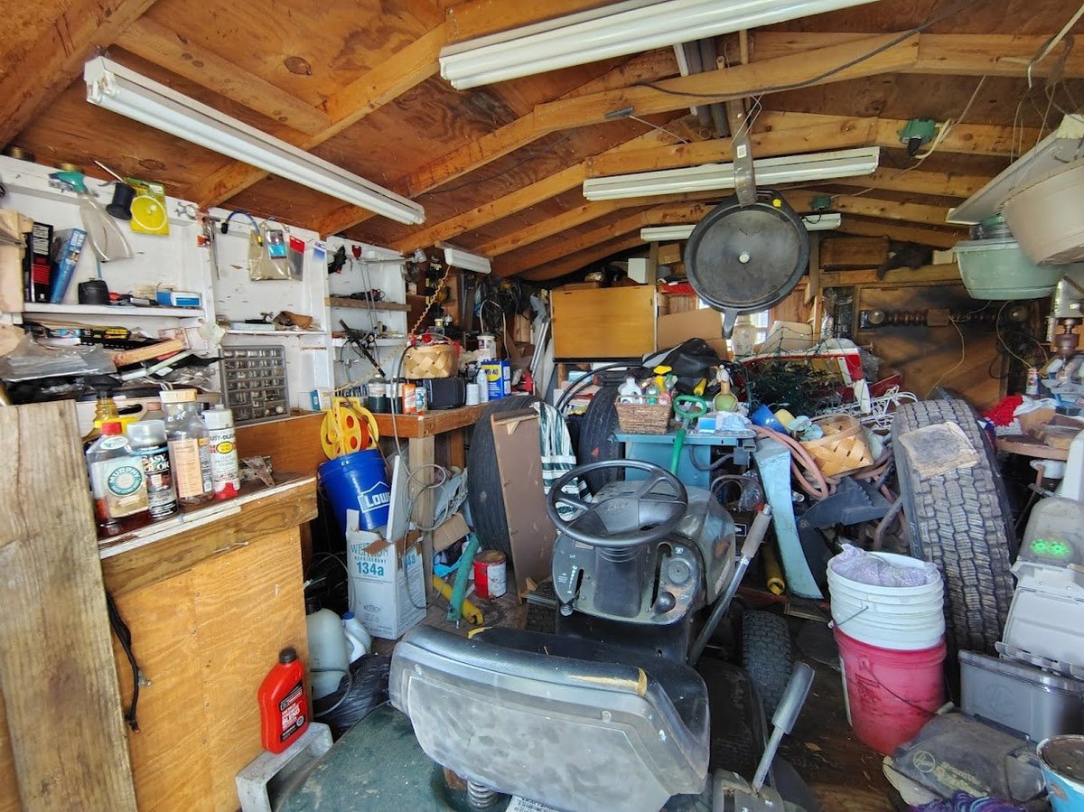 Cluttered garage workshop with wooden beams, shelving, tools, equipment, and storage containers throughout the space