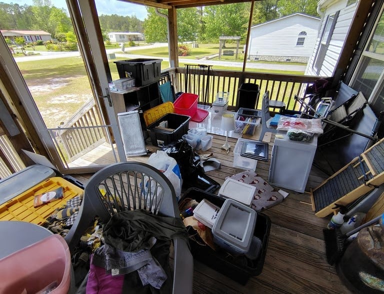 Cluttered screened porch with various boxes, storage items, and furniture overlooking a residential driveway