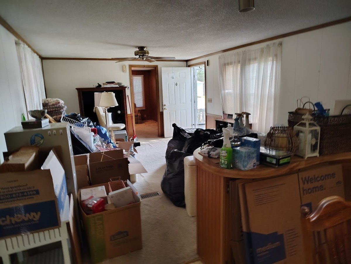 Cluttered room filled with cardboard boxes, furniture, and miscellaneous items during a move or storage situation.