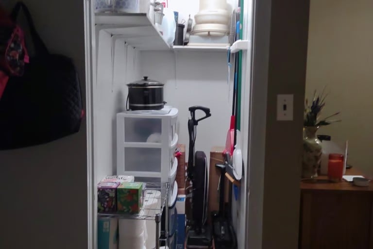 Open closet interior showing shelves with microwave, storage containers, cleaning equipment, and various household items organized on white shelving