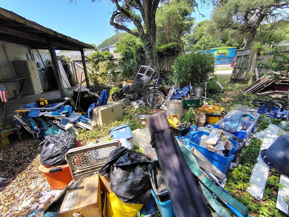 Cluttered backyard junkyard with scattered items, old furniture, bicycles, and miscellaneous junk piled throughout overgrown yard.